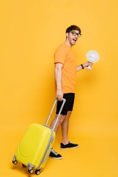 Homem animado em óculos com mala segurando ventilador elétrico no amarelo — Stock Photo