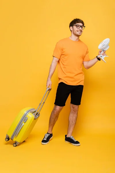 Sonriente hombre con bolsa de viaje sosteniendo ventilador eléctrico en amarillo - foto de stock