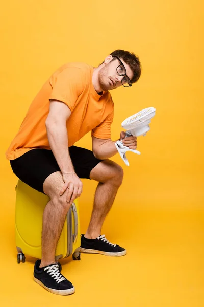 Frustrierter müder Mann leidet unter Hitze, während er einen elektrischen Ventilator hält und auf einem gelben Koffer sitzt — Stockfoto