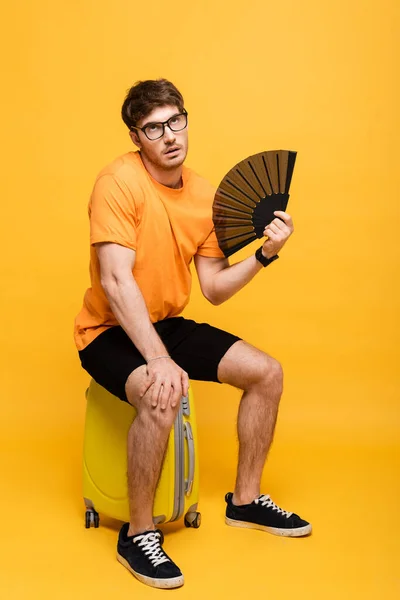 Frustrated man suffering from heat while using hand fan and sitting on suitcase on yellow — Stock Photo