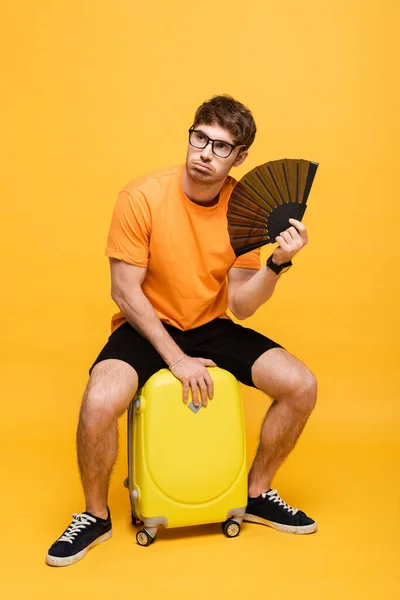 Confused man suffering from heat while using hand fan and sitting on suitcase on yellow — Stock Photo