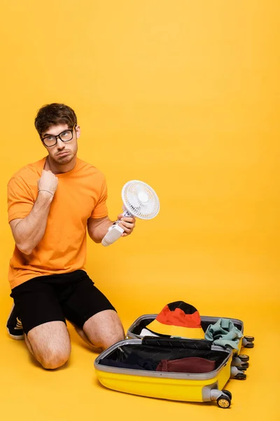 Homem cansado que embala o ventilador elétrico no saco de viagem no amarelo — Fotografia de Stock