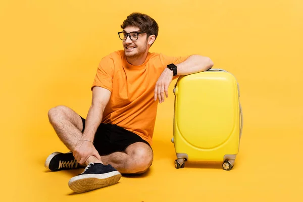 Happy tourist sitting near travel bag for summer vacation on yellow — Stock Photo