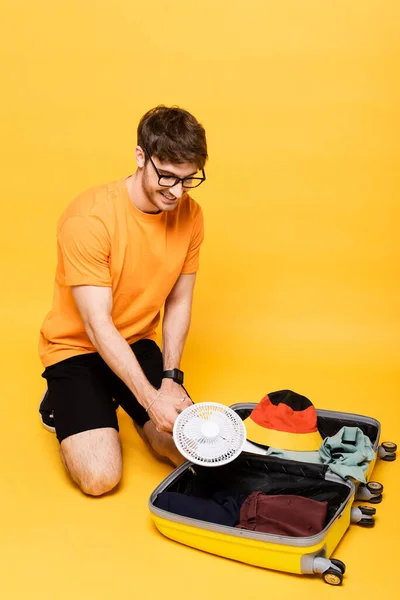 Happy man packing electric fan into travel bag on yellow — Stock Photo