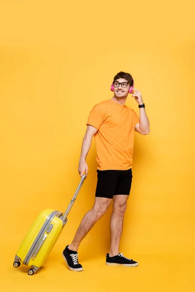 Счастливый красивый мужчина в наушниках ходит с дорожной сумкой на желтом — стоковое фото