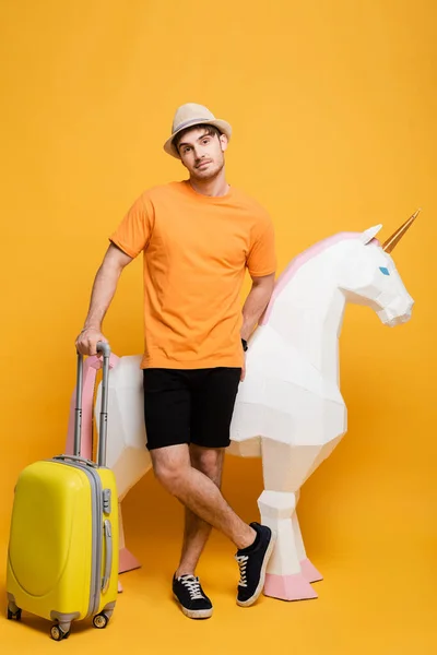 Young male traveler in hat standing with unicorn and suitcase on yellow — Stock Photo