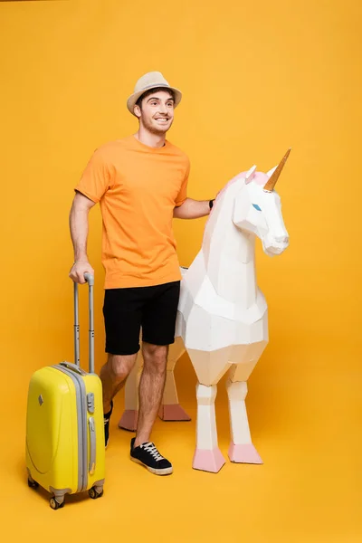 Smiling traveler in hat standing with unicorn and suitcase on yellow — Stock Photo