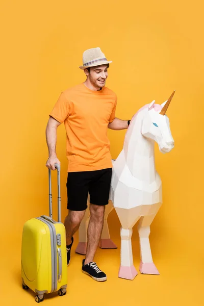 Cheerful traveler in hat standing with unicorn and suitcase on yellow — Stock Photo