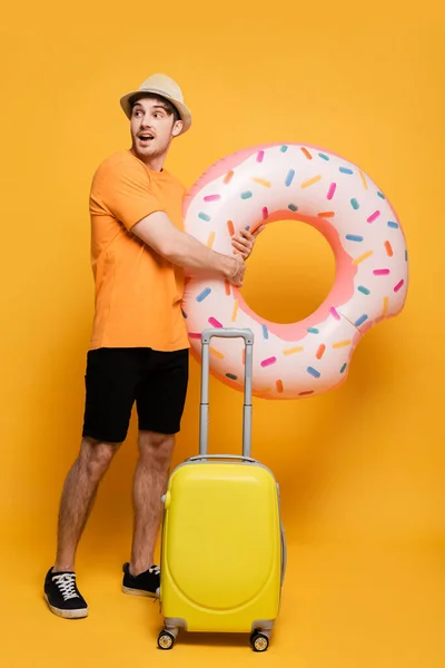 Молодий збуджений чоловік з валізою і надувний пончик готовий до літніх канікул на жовтому — стокове фото