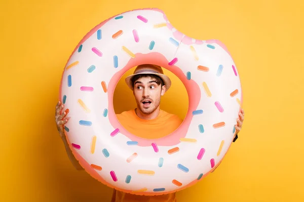 Hombre excitado con boca abierta de pie en sombrero con rosquilla inflable en amarillo - foto de stock