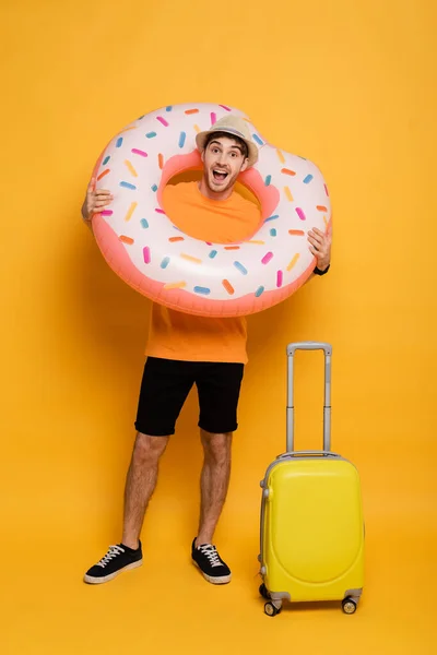 Excited man with suitcase and inflatable donut ready for summer vacation on yellow — Stock Photo
