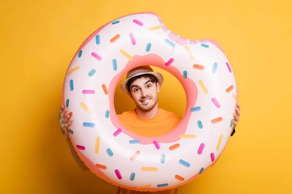 Fröhlicher Mann mit Hut steht mit aufblasbarem Donut auf gelb — Stockfoto