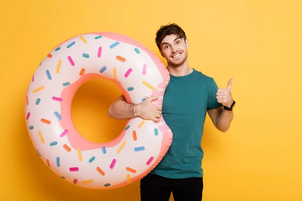 Fröhlicher Mann zeigt Daumen hoch und steht mit aufblasbarem Donut auf gelb — Stockfoto
