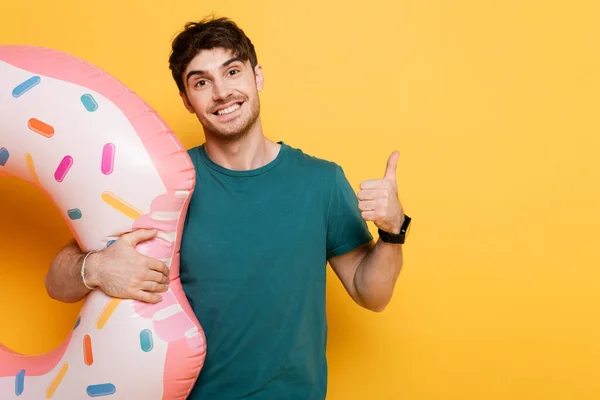 Sonriente hombre mostrando el pulgar hacia arriba mientras está de pie con rosquilla inflable en amarillo - foto de stock