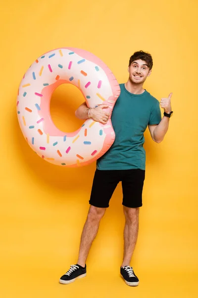 Glücklicher Mann zeigt Daumen nach oben, während er mit aufblasbarem Donut auf gelb steht — Stockfoto