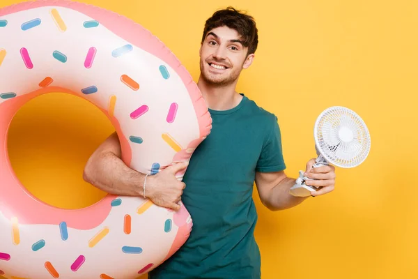 Homme souriant tenant donut gonflable et petit ventilateur électrique sur jaune — Photo de stock