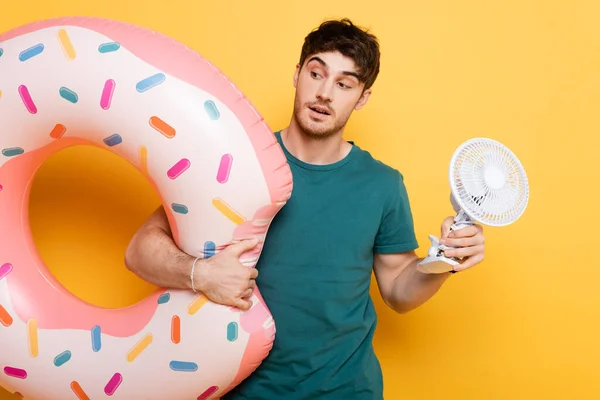 Überraschter Mann mit aufblasbarem Donut und kleinem Elektroventilator auf gelb — Stockfoto