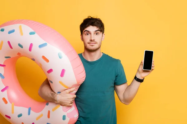 Junger Mann mit aufblasbarem Donut hält Smartphone mit leerem Bildschirm auf gelb — Stockfoto