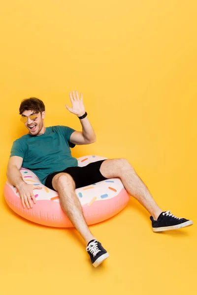 Hombre excitado en gafas de sol agitando la mano y sentado en donut inflable en amarillo - foto de stock