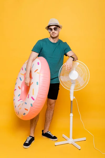 Schöner Mann mit Sonnenbrille und Hut hält aufblasbaren Donut in der Nähe von elektrischen Ventilator auf gelb — Stockfoto