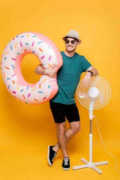Uomo sorridente in occhiali da sole e cappello tenendo ciambella gonfiabile mentre in piedi vicino ventilatore elettrico sul giallo — Foto stock
