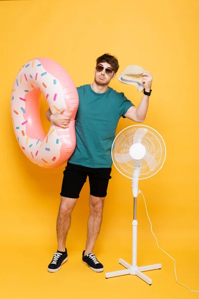 Homme confus dans les lunettes de soleil tenant chapeau et beignet gonflable tout en se tenant près ventilateur électrique sur jaune — Photo de stock