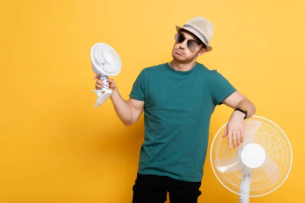 Beau jeune homme en lunettes de soleil et chapeau debout avec deux ventilateurs électriques sur jaune — Photo de stock