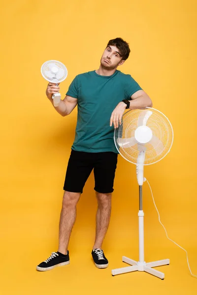 Homem confuso de pé com dois ventiladores elétricos no amarelo — Fotografia de Stock