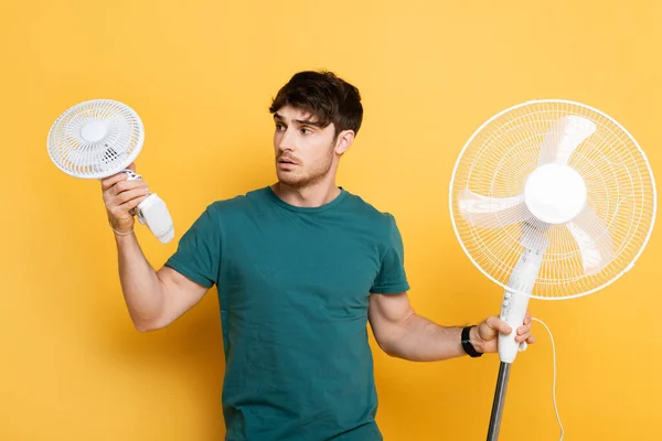 Jeune homme frustré tenant avec deux ventilateurs électriques sur jaune — Photo de stock