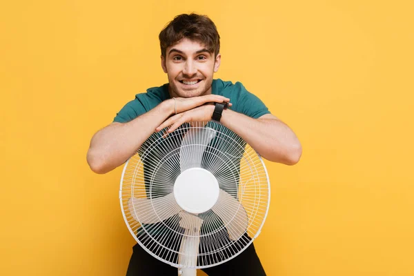 Lächelnder junger Mann steht mit elektrischem Ventilator auf gelb — Stockfoto