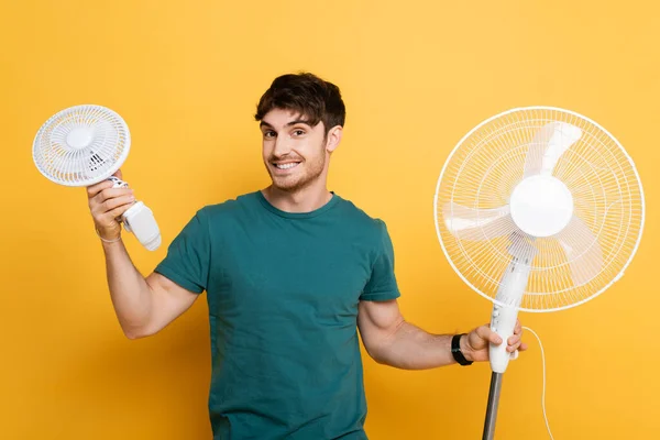 Homme heureux debout avec deux ventilateurs électriques sur jaune — Photo de stock