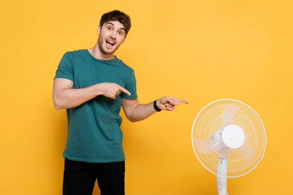 Jovem alegre apontando com os dedos para o ventilador elétrico no amarelo — Fotografia de Stock