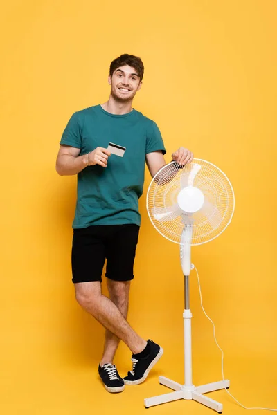 Sorridente jovem segurando cartão de crédito, enquanto de pé com ventilador elétrico no amarelo — Fotografia de Stock