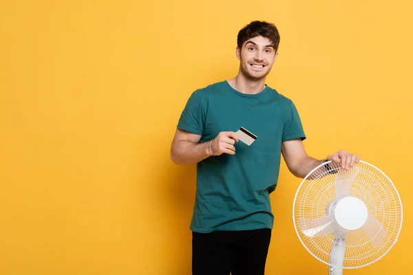 Homme heureux tenant carte de crédit tout en se tenant debout avec ventilateur électrique sur jaune — Photo de stock