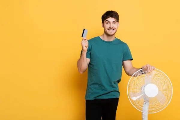 Uomo sorridente in possesso di carta di credito in piedi con ventilatore elettrico sul giallo — Foto stock