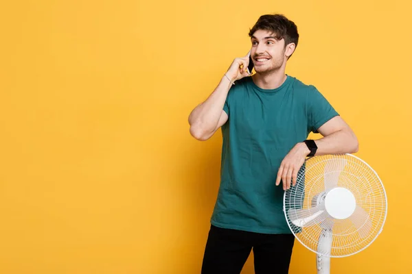 Smiling man talking on cellphone while standing near electric fan on yellow — Stock Photo