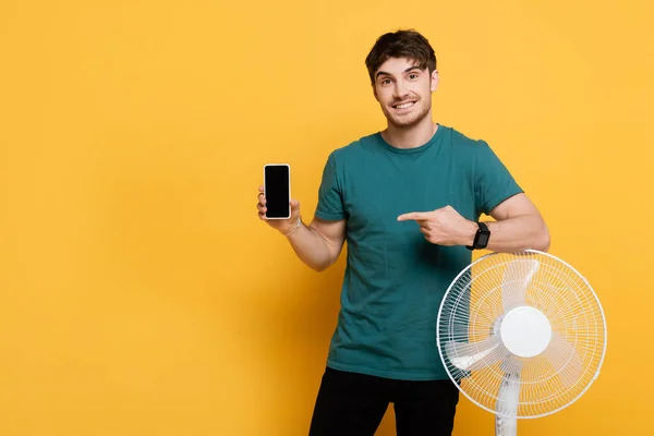 Lächelnder Mann zeigt auf Smartphone mit leerem Bildschirm, während er neben Elektroventilator auf gelb steht — Stockfoto