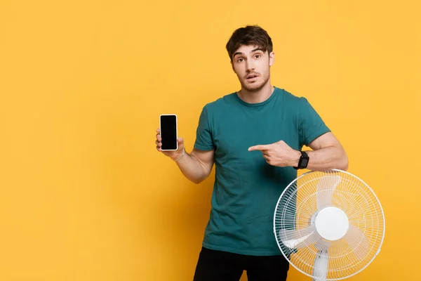 Schockierter Mann zeigt mit Finger auf Smartphone mit leerem Bildschirm, während er neben Elektroventilator auf gelb steht — Stockfoto