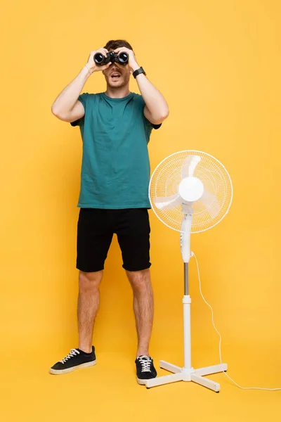 Jeune homme regardant à travers les jumelles tout en se tenant debout avec ventilateur électrique sur jaune — Photo de stock