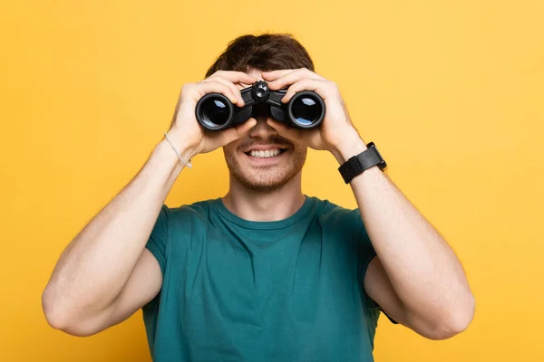 Cheerful man looking through binoculars on yellow — Stock Photo