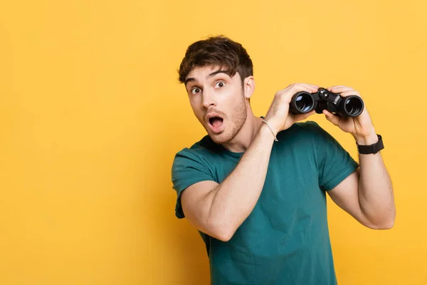 Beau homme surpris tenant des jumelles et regardant la caméra sur jaune — Photo de stock