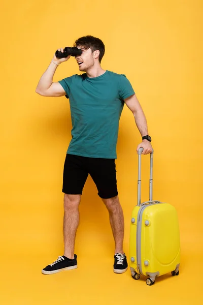 Збуджений чоловік з валізою готовий до літніх канікул і дивиться через бінокль на жовтому — стокове фото