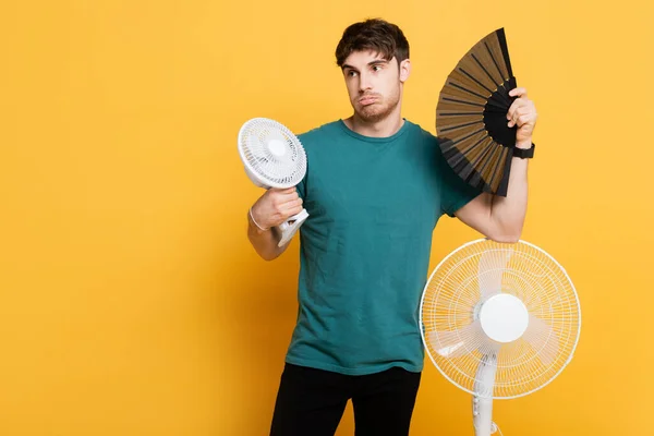 Confused man suffering from heat with two electric fans and hand fan on yellow — Stock Photo