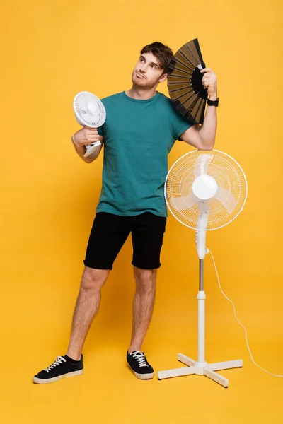 Schöner Mann mit zwei elektrischen Ventilatoren und Handventilator auf gelb — Stockfoto