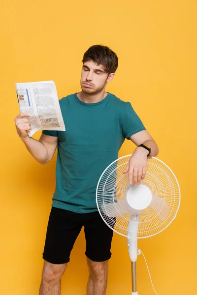 Young man suffering from heat with electric fan and newspaper as hand fan on yellow — Stock Photo