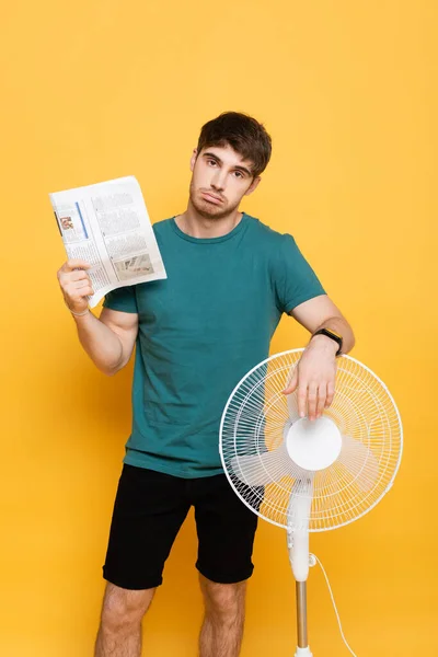 Homem triste que sofre de calor com ventilador elétrico e jornal como ventilador de mão no amarelo — Fotografia de Stock