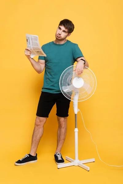 Confused man suffering from heat with electric fan and newspaper as hand fan on yellow — Stock Photo