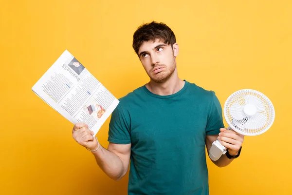 Frustrated young man suffering from heat with electric fan and newspaper as hand fan on yellow — Stock Photo