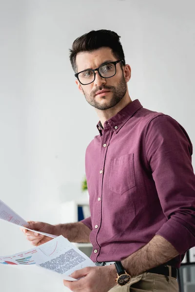 Junger, seriöser Geschäftsmann mit Brille schaut weg, während er Dokumente in der Hand hält — Stockfoto