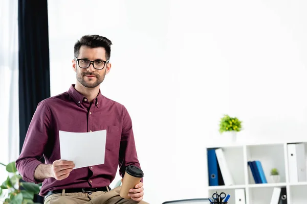 Sonriente, guapo hombre de negocios sosteniendo documento y café para ir mientras mira a la cámara - foto de stock